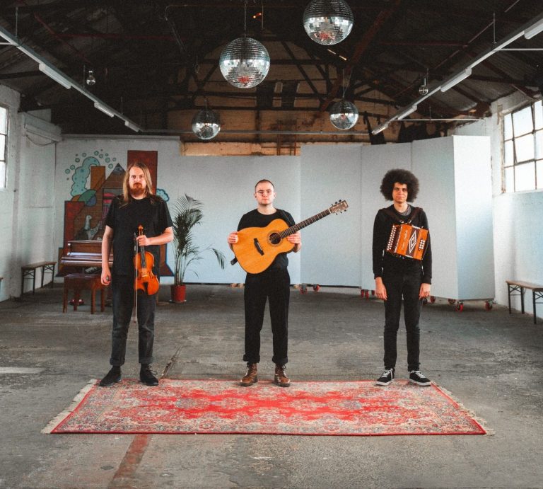 Three men from granny's attic stand in a warehouse on a rug with instruments